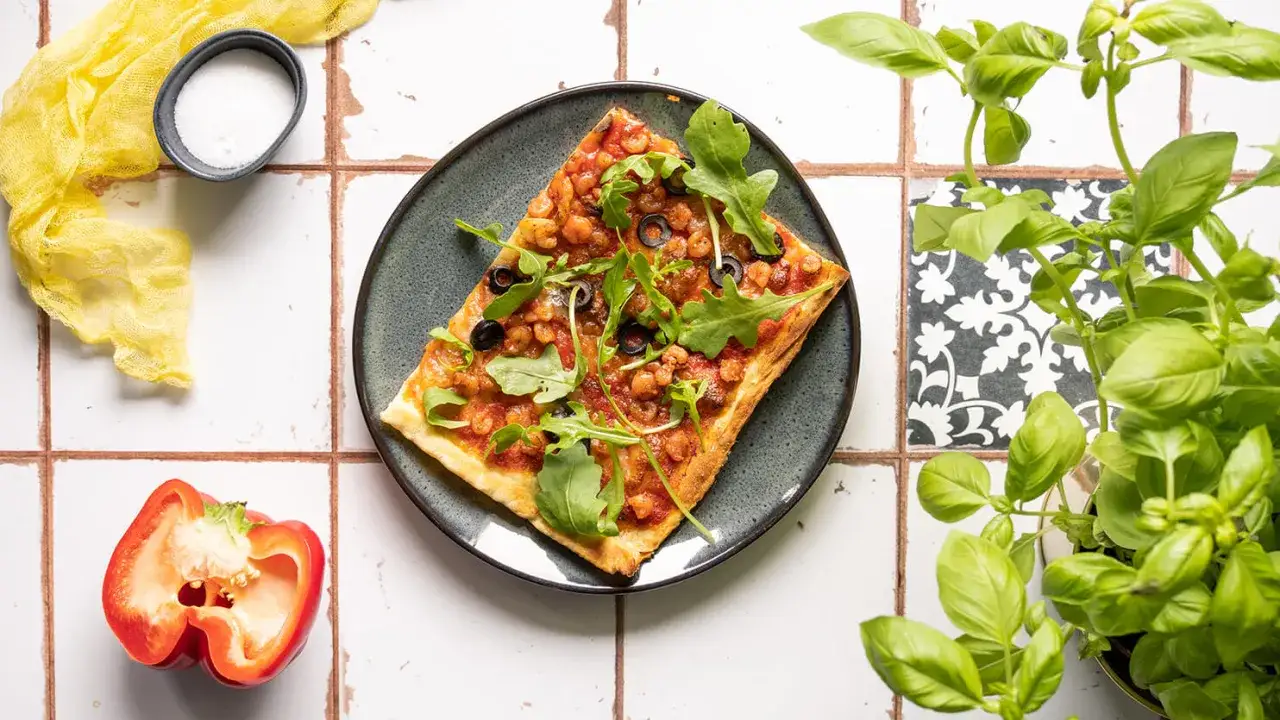 Aromatyczna pizza z krewetkami, rukolą i oliwkami na talerzu, obok świeża bazylia i papryka.