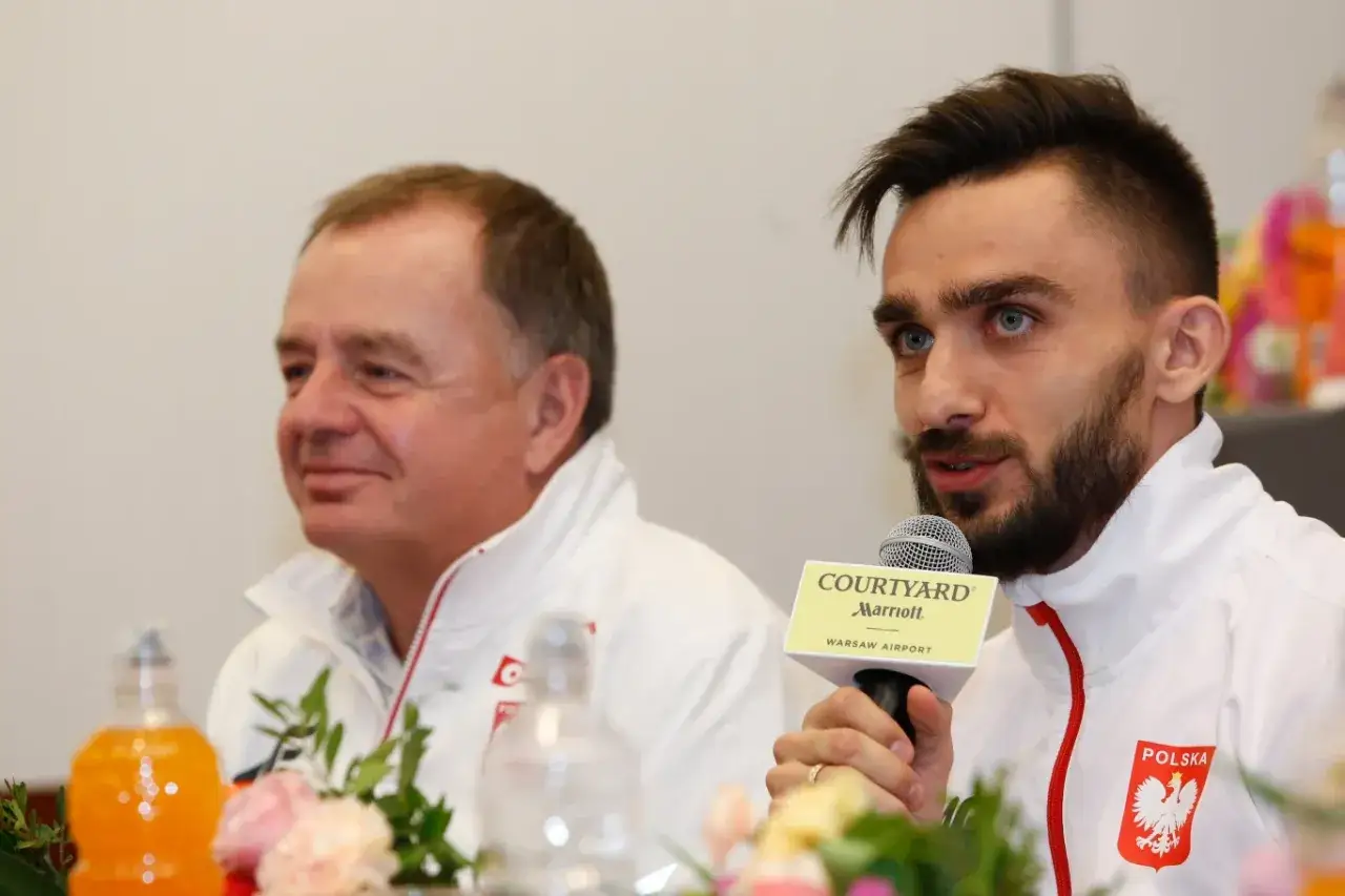 Zbigniew Król, trener, z uśmiechem na twarzy podczas konferencji prasowej. Obok siedzi młodszy mężczyzna z mikrofonem.
