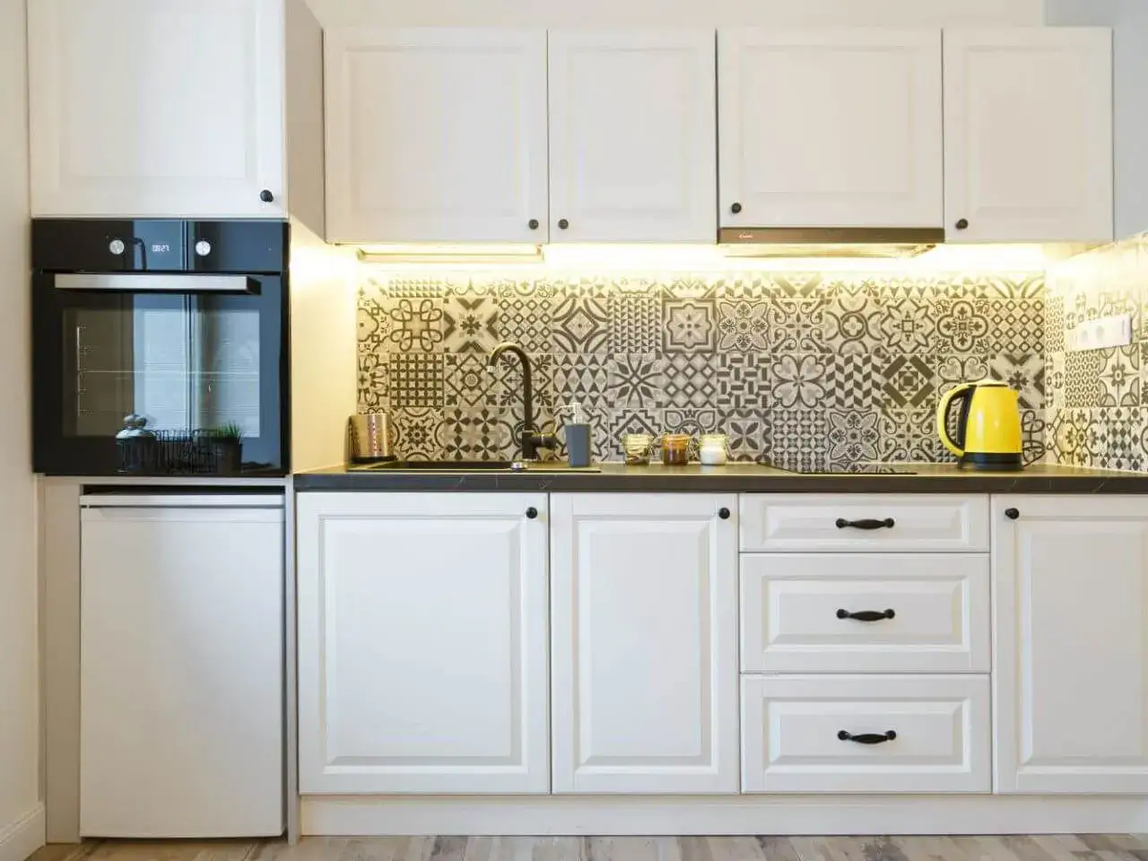 Nowoczesna kuchnia z białymi szafkami i efektownymi płytkami we wzory marokańskie nad blatem. Obok piekarnika stoi ż&oacute;łty czajnik.