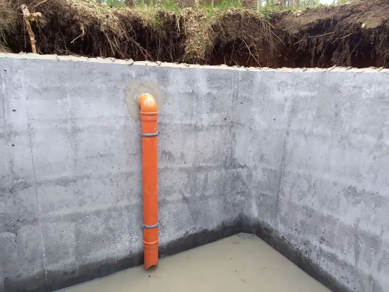 Pomarańczowa rura do szamba w betonowym ogrodzeniu, z widoczną wodą na dnie.