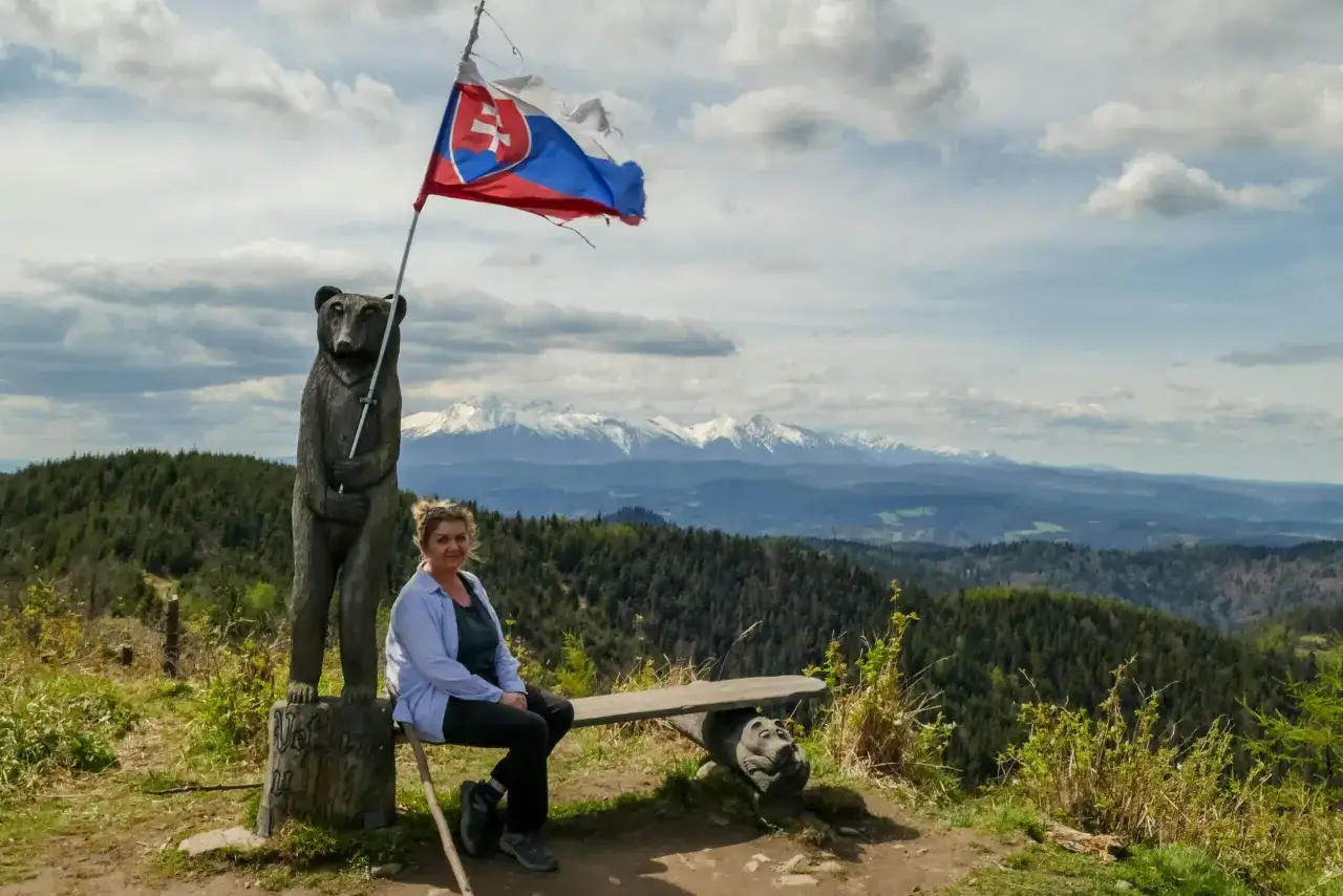 Kobieta siedzi na ławce pod słowacką flagą, obok rzeźby niedźwiedzia. Widok na g&oacute;ry z **veterny vrch**.