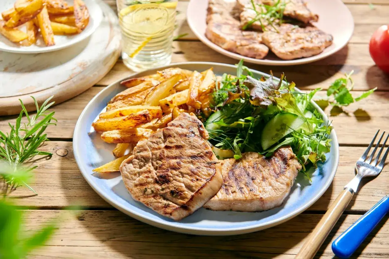 Soczysty schab z grilla podany z chrupiącymi frytkami i świeżą sałatką. Idealny letni posiłek na drewnianym stole.