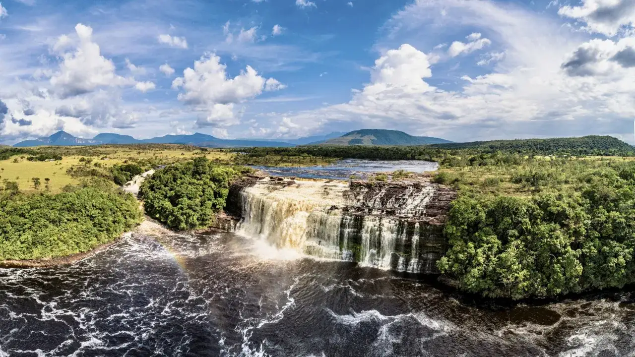 Wodospad w Parku Narodowym Canaima, z tęczą w spienionej wodzie, otoczony bujną zielenią i górami w tle.