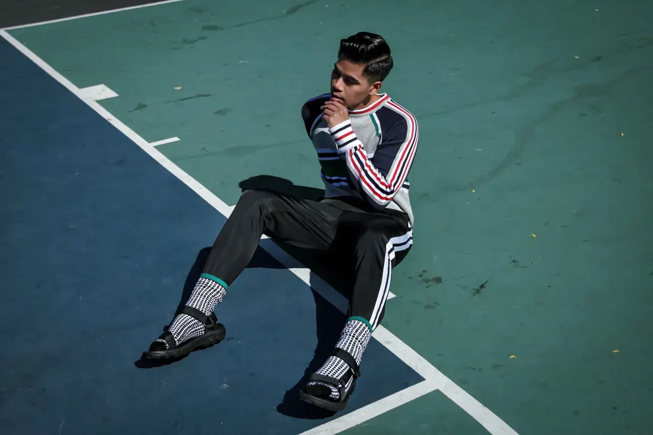 Młody mężczyzna w stylowym swetrze i spodniach siedzi na boisku. Jego stopy zdobią czarne sandały i biało-czarne skarpety w kratkę.