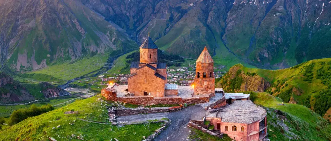 Kości&oacute;ł Gergeti Tr&oacute;jcy Świętej na tle g&oacute;r. Wioska w dolinie. Ceny w Gruzji mogą być zaskakująco przystępne.