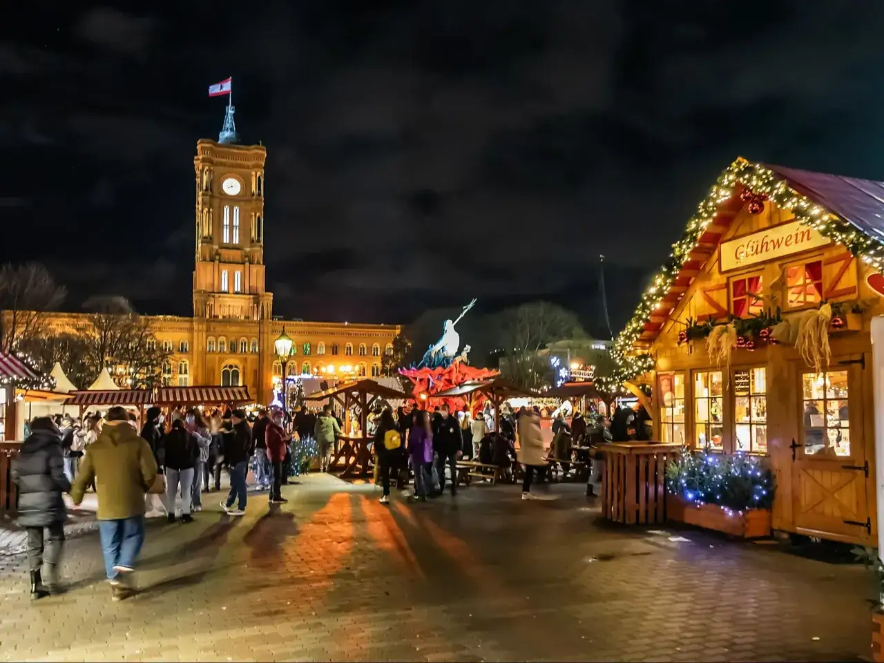 Tłoczny jarmark bożonarodzeniowy w Berlinie nocą. W tle Rotes Rathaus, a po prawej drewniana budka z napisem 