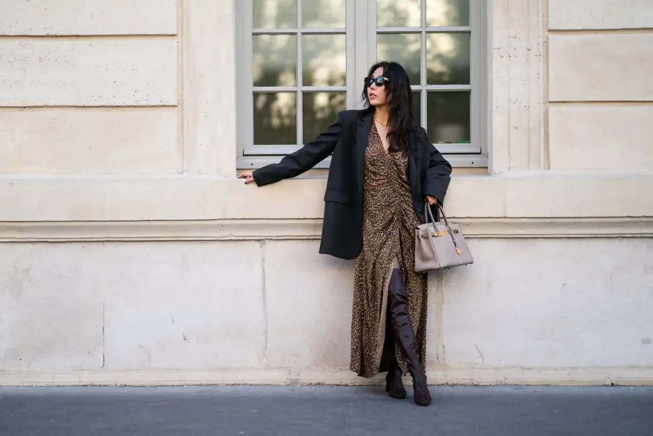 Elegancka kobieta w sukience w panterkę i czarnym blazerze, z beżową torebką. Długie, brązowe kozaki dla niskiej osoby dodają jej wzrostu i stylu.