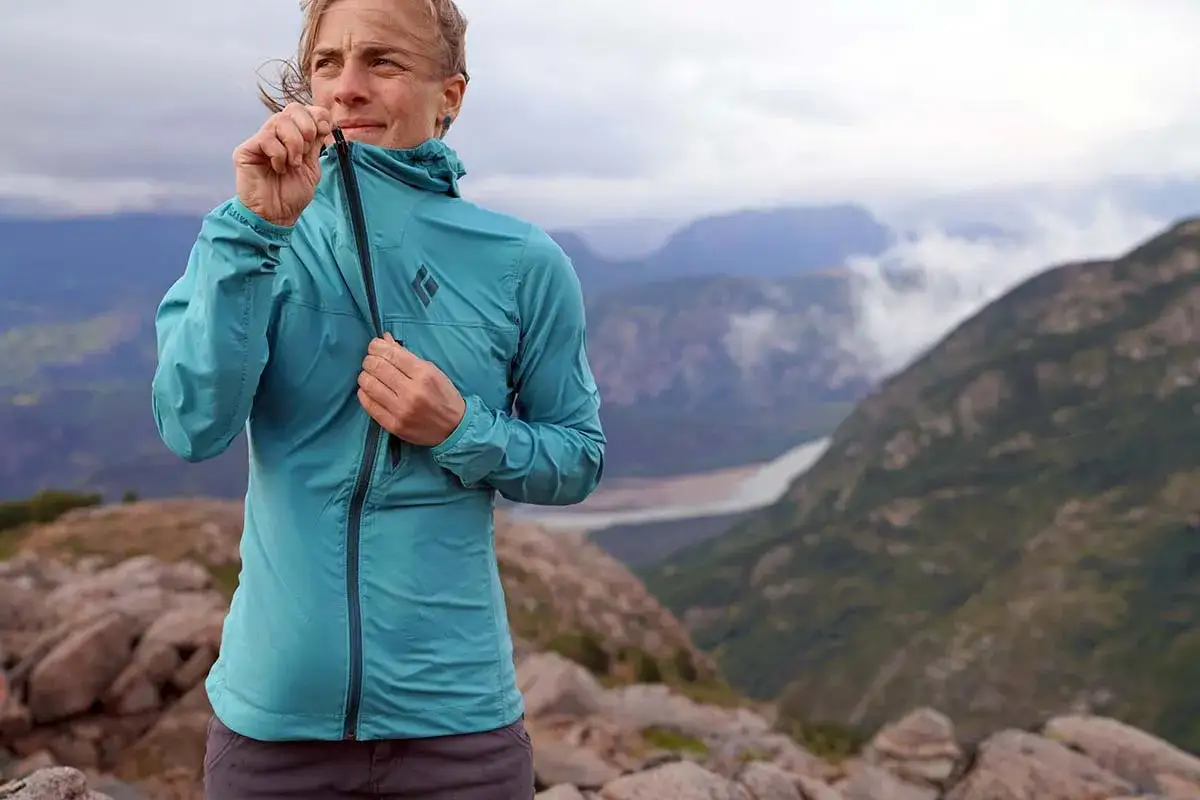 Kobieta zapina turkusową kurtkę softshell, idealną na górskie wędrówki. Wiatr rozwiewa jej włosy, a w tle widać mgliste szczyty.