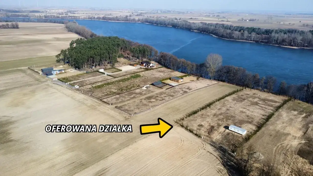 Działka z linią brzegową jeziora, otoczona drzewami i polami. Widać kilka budynków i drogę.