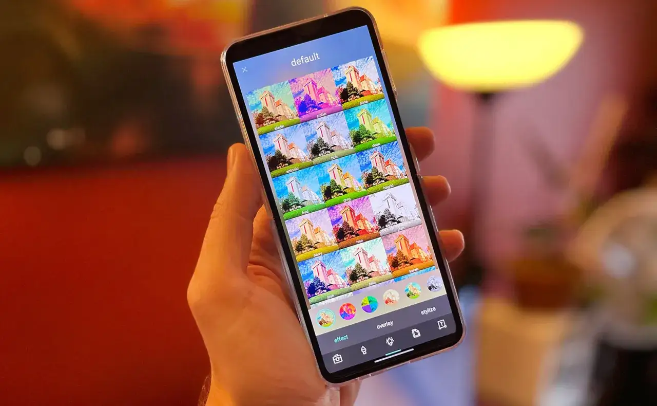 Dłoń trzyma smartfon z otwartym programem do obr&oacute;bki zdjęć na telefon. Widoczne są liczne filtry z podglądem efektu na zdjęciu zamku.