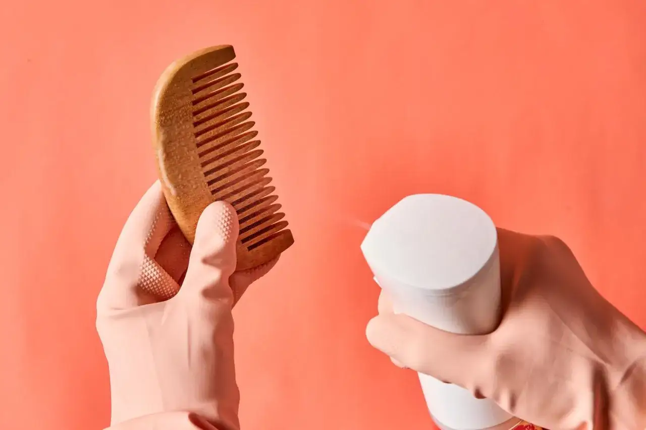 Rękawiczka w r&oacute;żowym kolorze trzyma drewniany grzebień, a druga rękawiczka rozpyla na niego płyn. Dowiedz się, jak wyczyścić szczotkę do włos&oacute;w.