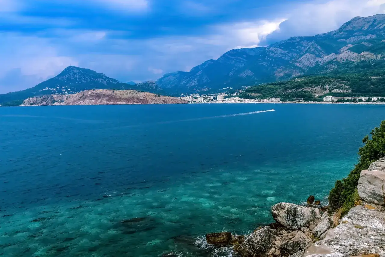 Widok na turkusowe morze i g&oacute;ry w Sutomore. Widoczne białe budynki kurortu i ł&oacute;dź na wodzie. To idealne miejsce na odkrywanie sutomore atrakcje.