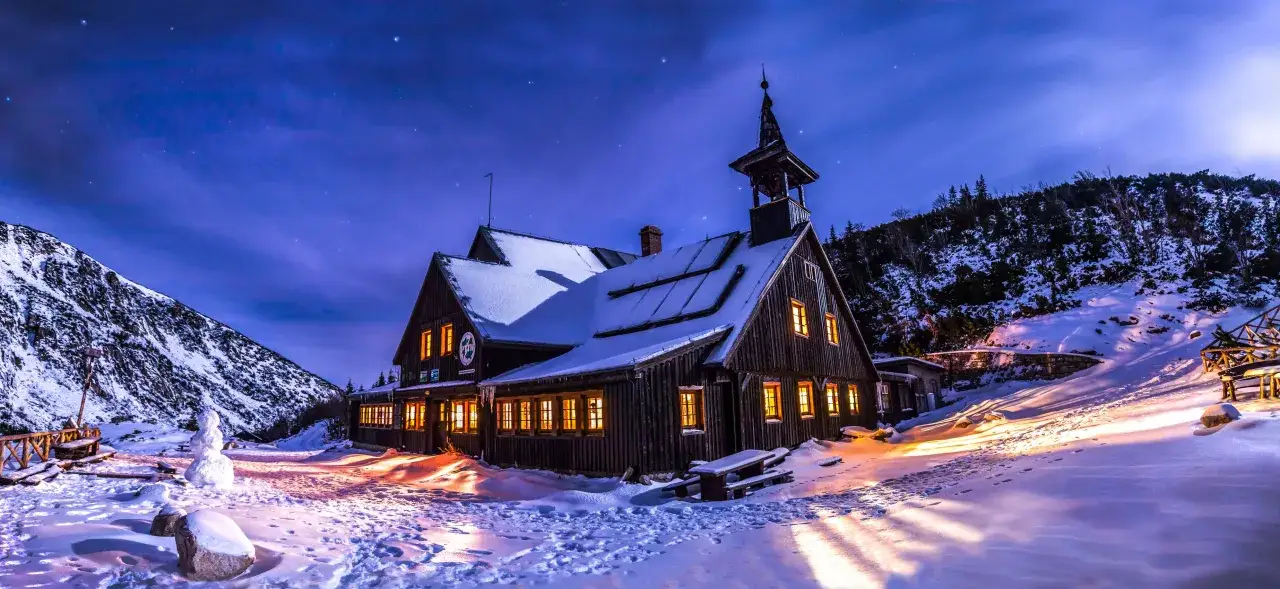 Drewniane schronisko oświetlone ciepłym światłem, otoczone śniegiem i nocnym niebem pełnym gwiazd. Idealne miejsce, gdzie na ferie zimowe w g&oacute;ry.
