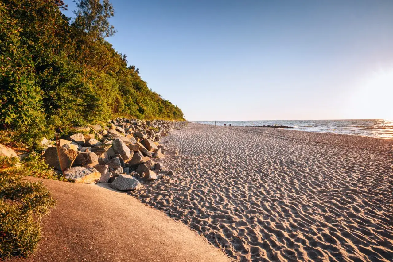 Plaża nad Bałtykiem, gdzie jest najwięcej jodu w Polsce. Słońce zachodzi nad spokojnym morzem, a piasek jest usiany kamieniami i zielenią.