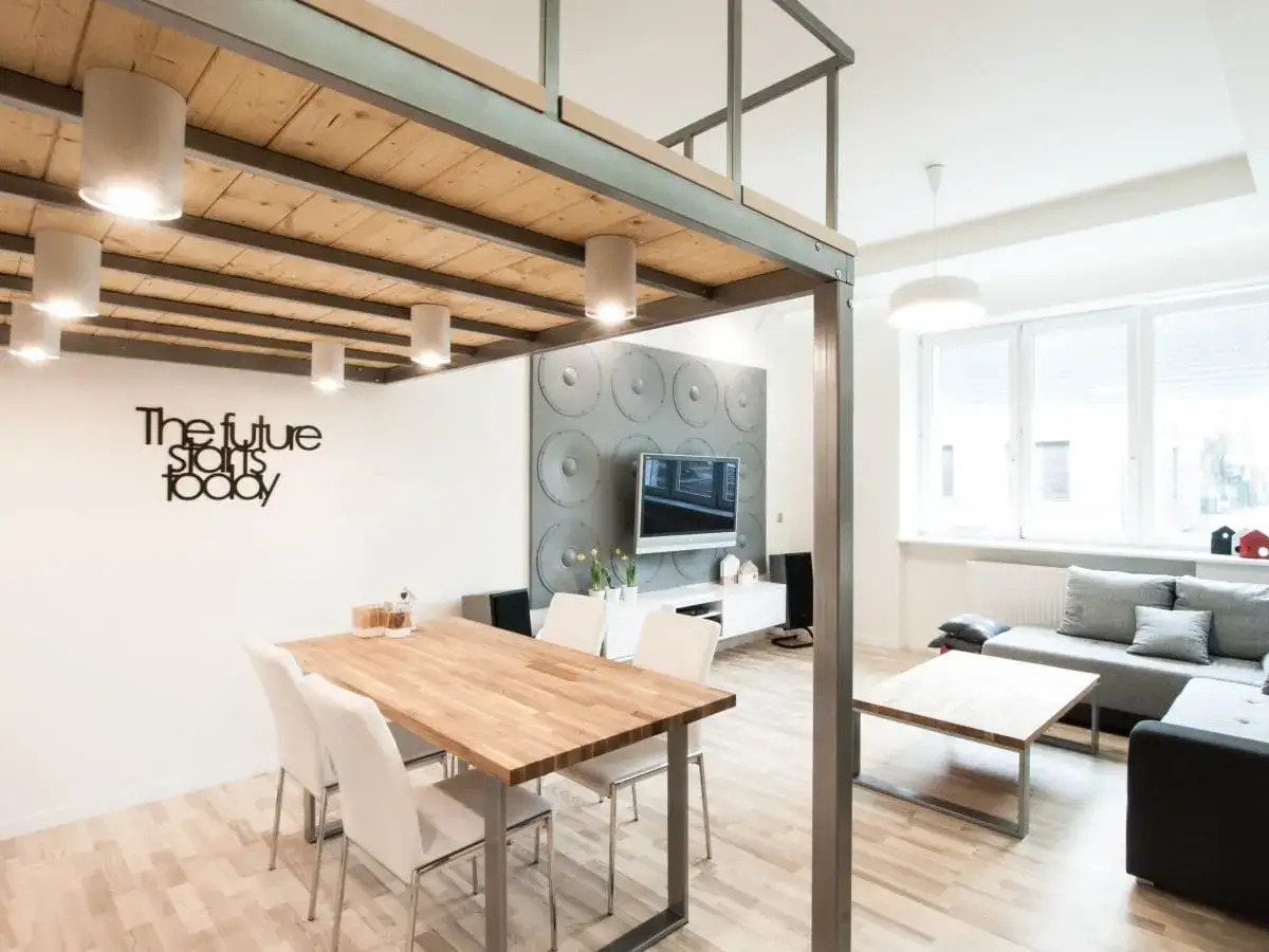 Nowoczesne mieszkanie z antresolą, jadalnia z drewnianym stołem i białe krzesła. W tle salon z szarą kanapą i stolikiem kawowym.