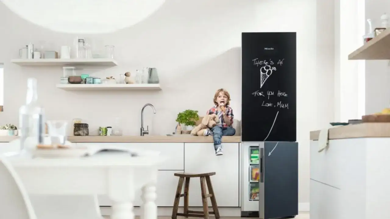 Chłopiec z pluszowym psem siedzi na blacie w kuchni, jedząc lody. Obok lod&oacute;wka z tablicą, na kt&oacute;rej mama napisała 