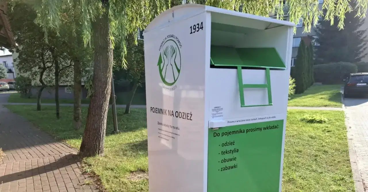 Białe kontenery na ubrania z zielonymi elementami stoją na chodniku obok drzew.