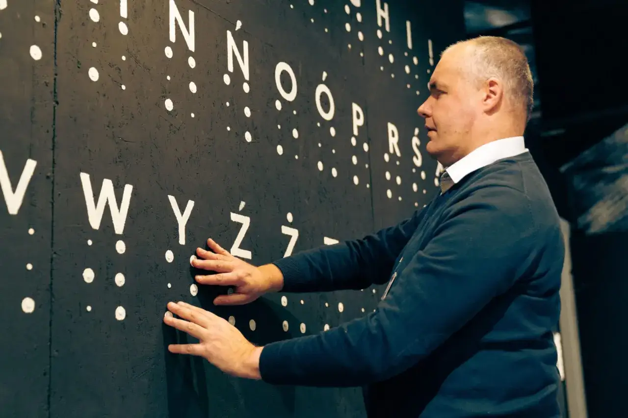 Mężczyzna dotyka wypukłych liter i kropek na czarnej ścianie. To wystawa w ciemności, która pozwala doświadczyć świata zmysłem dotyku.