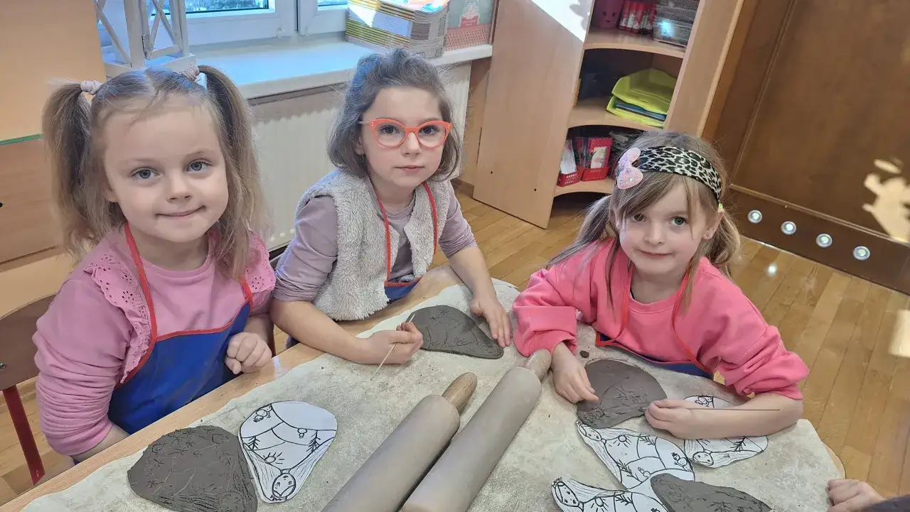 Trzy dziewczynki tworzą dzieła z gliny podczas warsztat&oacute;w. Na stole leżą wałki i gliniane formy.