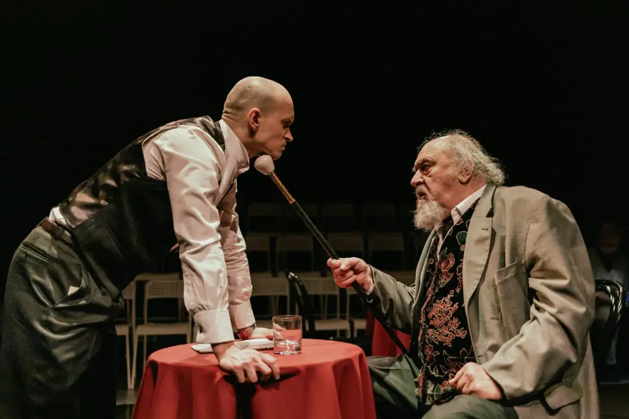 Dwóch aktorów na scenie, jeden pochylony nad stołem, drugi siedzi. Czy to scena z przedstawienia, które pomoże zrozumieć, ile zarabiają aktorzy w Polsce?