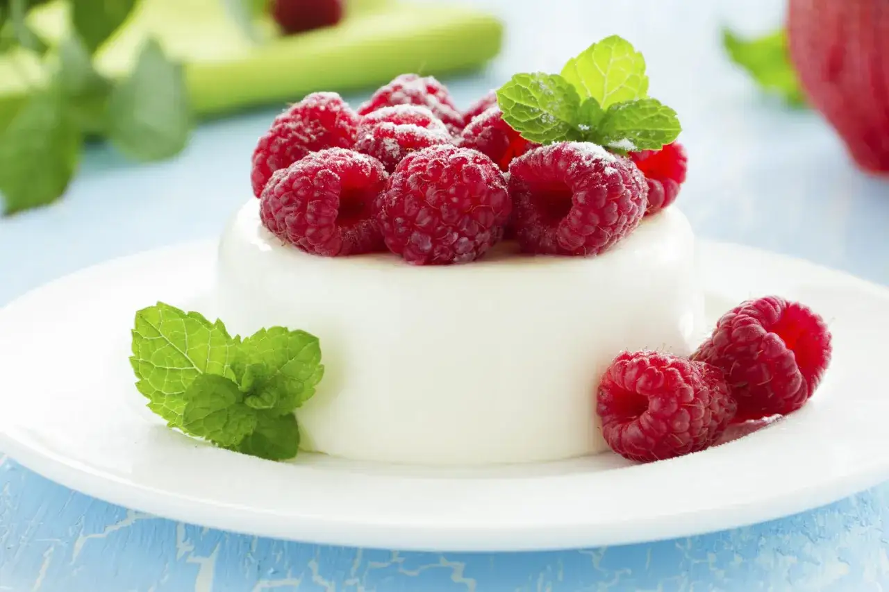 Pyszna panna cotta z malinami i miętą, posypana cukrem pudrem, prezentuje się apetycznie na białym talerzyku.