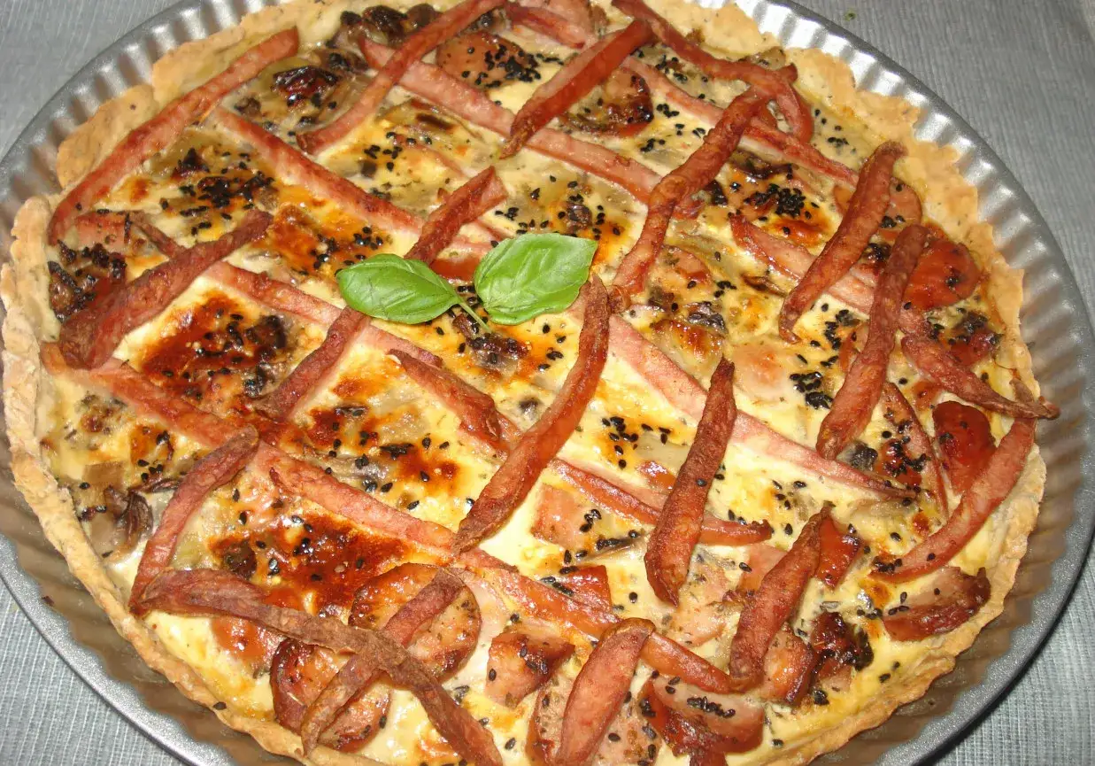 Pyszna tarta z kiełbasą, jajkami i serem, ozdobiona czarnym sezamem i listkiem bazylii.