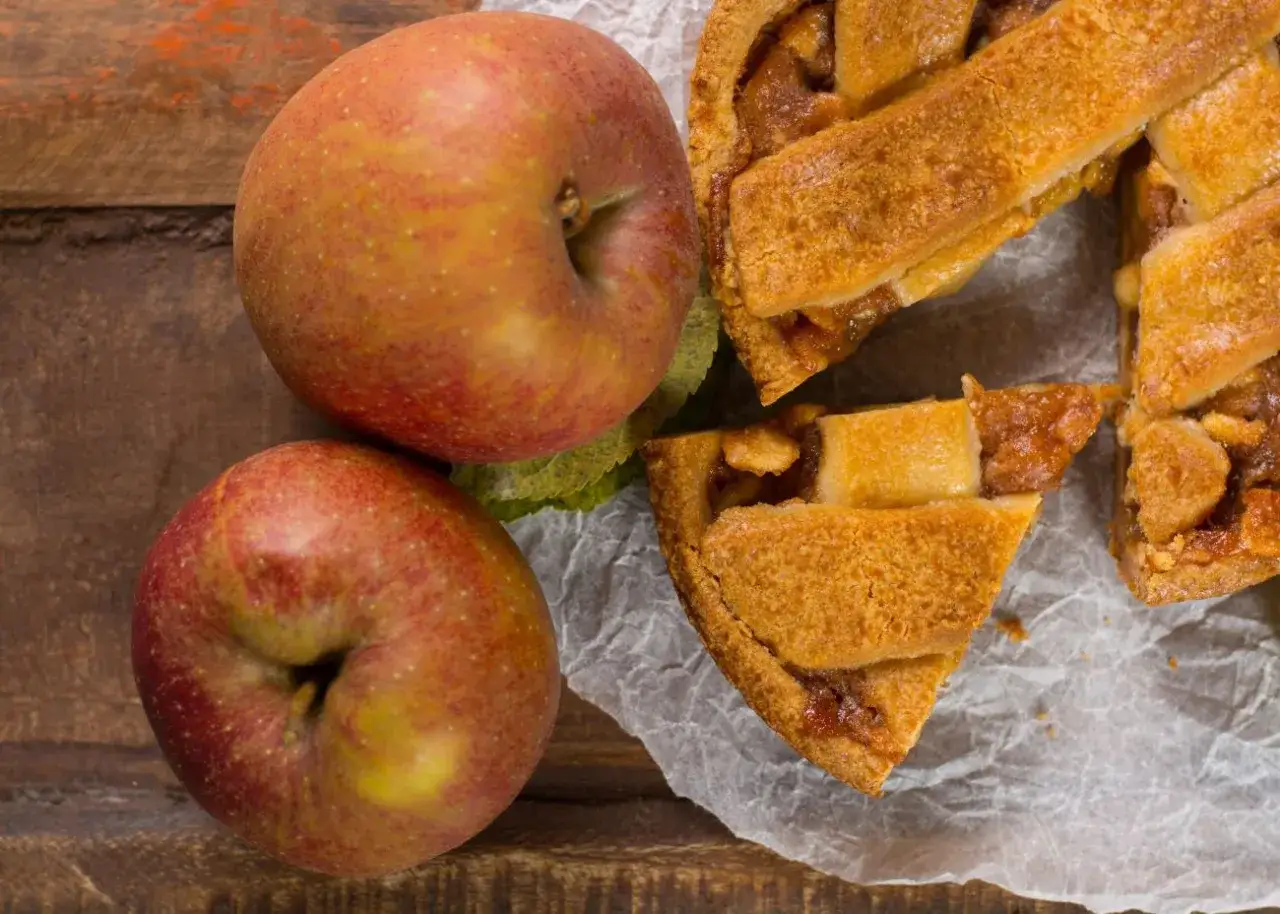 Dwie jabłka obok pysznej szarlotki. Zastanawiasz się, ile czasu piec szarlotkę, by była tak idealna?