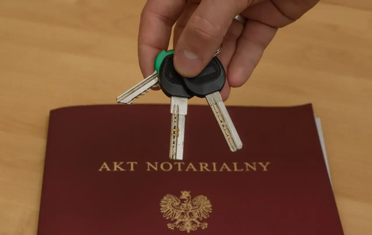 Dłoń trzyma klucze nad aktem notarialnym, symbolizującym akt własności domu.