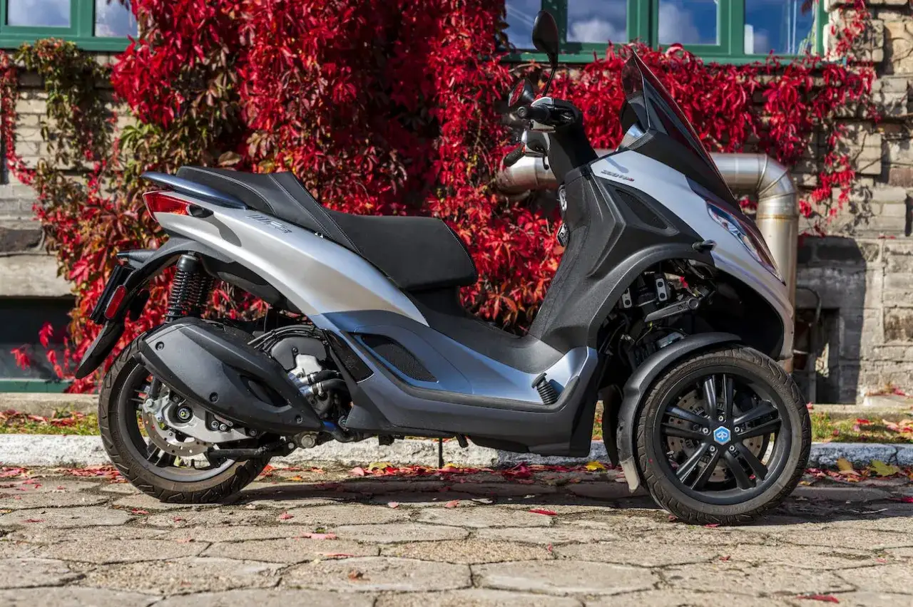Srebrny Piaggio MP3 300, tr&oacute;jkołowy skuter, prezentuje swoje **dane techniczne** na tle jesiennych liści i ceglanej ściany.