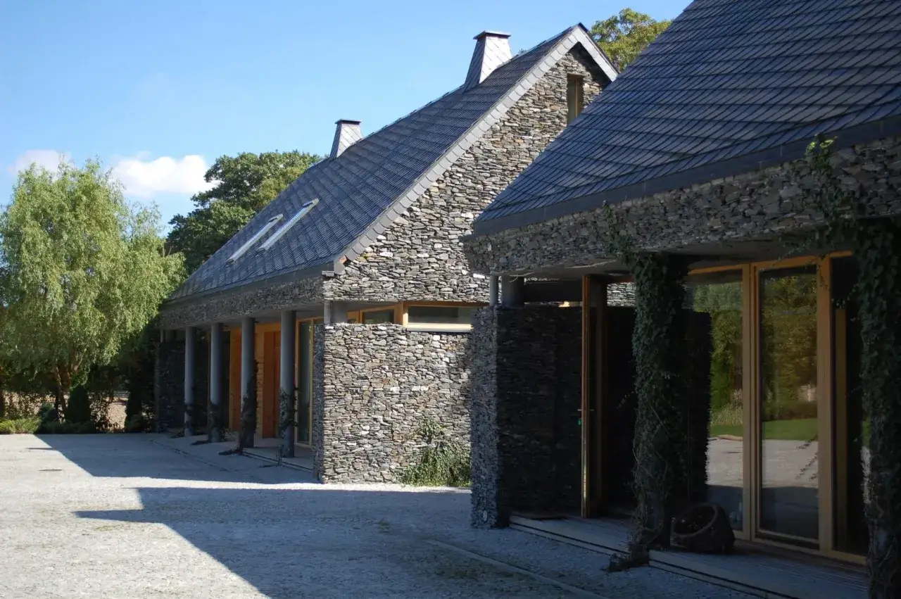 Nowoczesny dom z piaskowca, z kamiennymi ścianami i ciemnym dachem, otoczony zielenią.