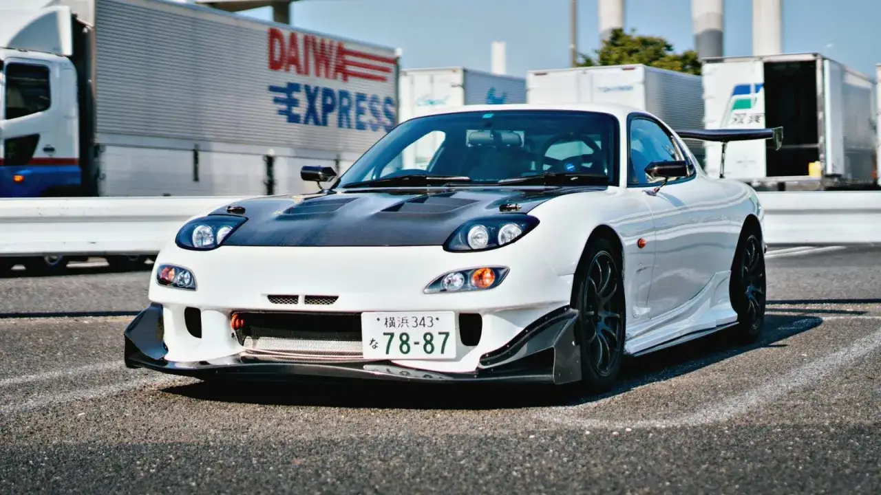 Biały, zmodyfikowany Mazda RX-7 z czarną maską i spojlerem, jeden z ikonicznych japońskich samochodów sportowych, zaparkowany na tle ciężarówek.