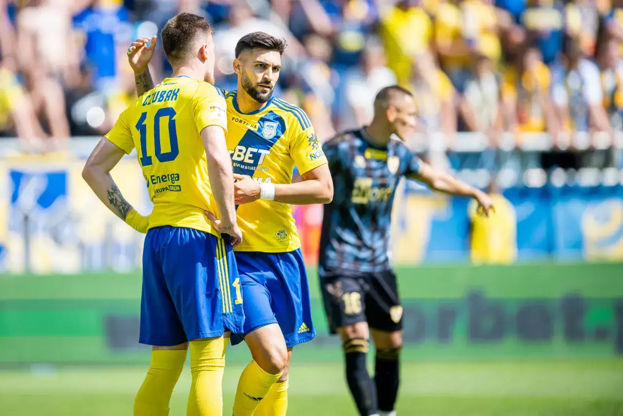 Piłkarze w żółtych strojach rozmawiają na boisku, być może omawiając, ile drużyn awansuje do ekstraklasy.