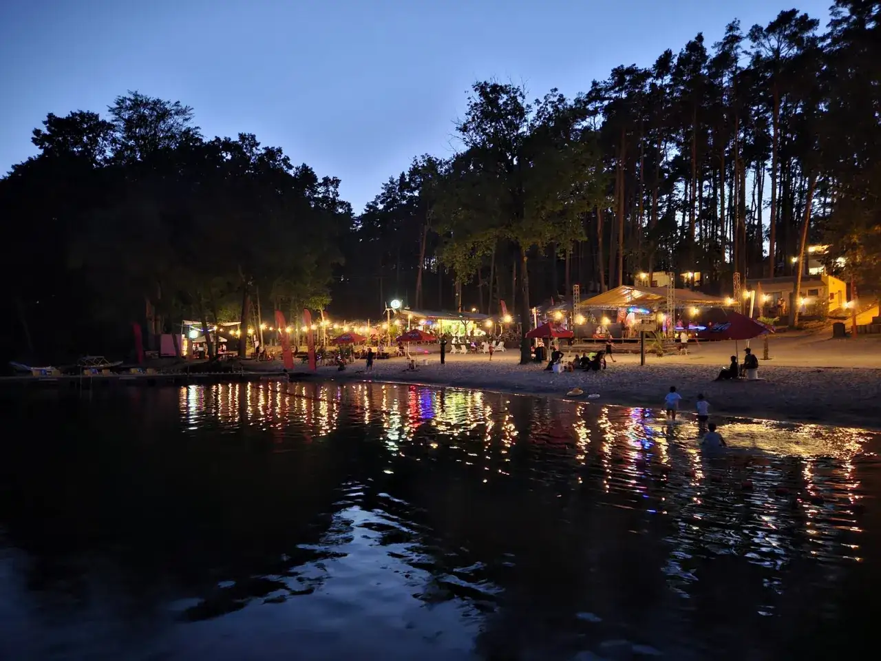 Wieczorny, magiczny camping Lubniewice nad jeziorem. Oświetlona plaża z ludźmi, parasolami i budynkami, odbijająca się w wodzie.