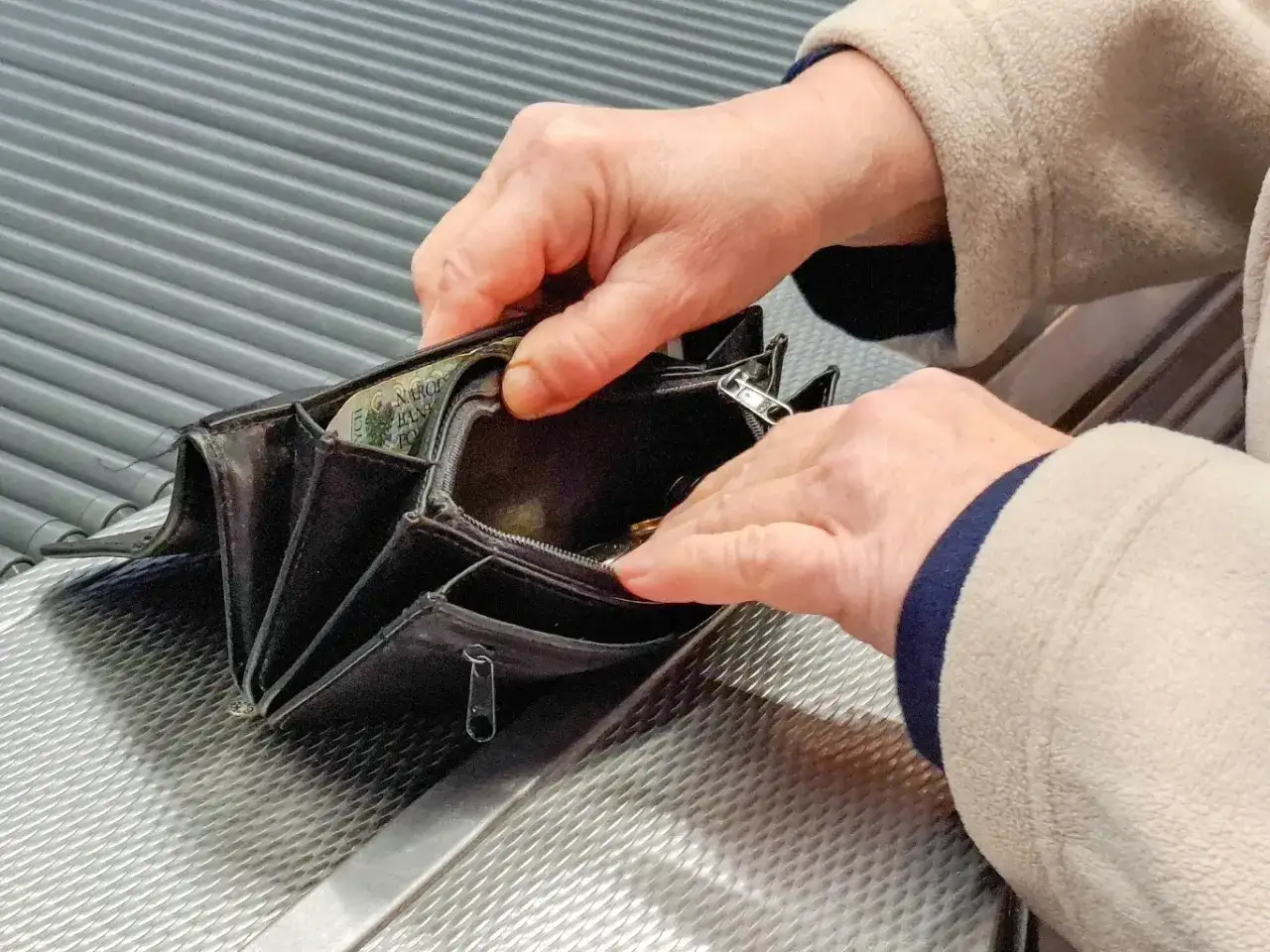 Dłonie starszej osoby przeglądają portfel, być może kiedy listonosz przynosi emeryturę. Widać kilka monet i banknot.