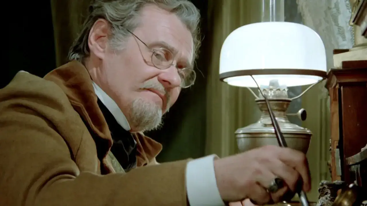 Starszy mężczyzna w okularach pochyla się nad biurkiem, pisząc przy lampie. Jego narracja w lalce tworzy atmosferę tajemnicy.