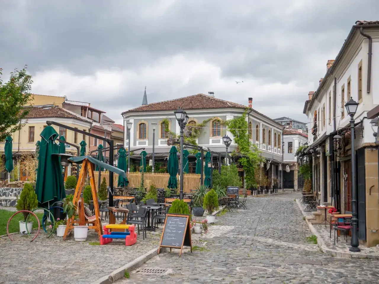 Urokliwy zaułek w Korce, Albania, z kawiarnianymi stolikami i zielonymi parasolami.