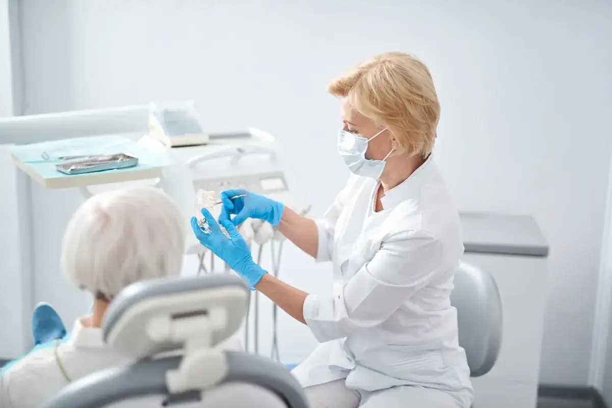 Dentystka w masce i rękawiczkach pokazuje pacjentowi model szczęki, omawiając usunięcie torbieli zęba NFZ.