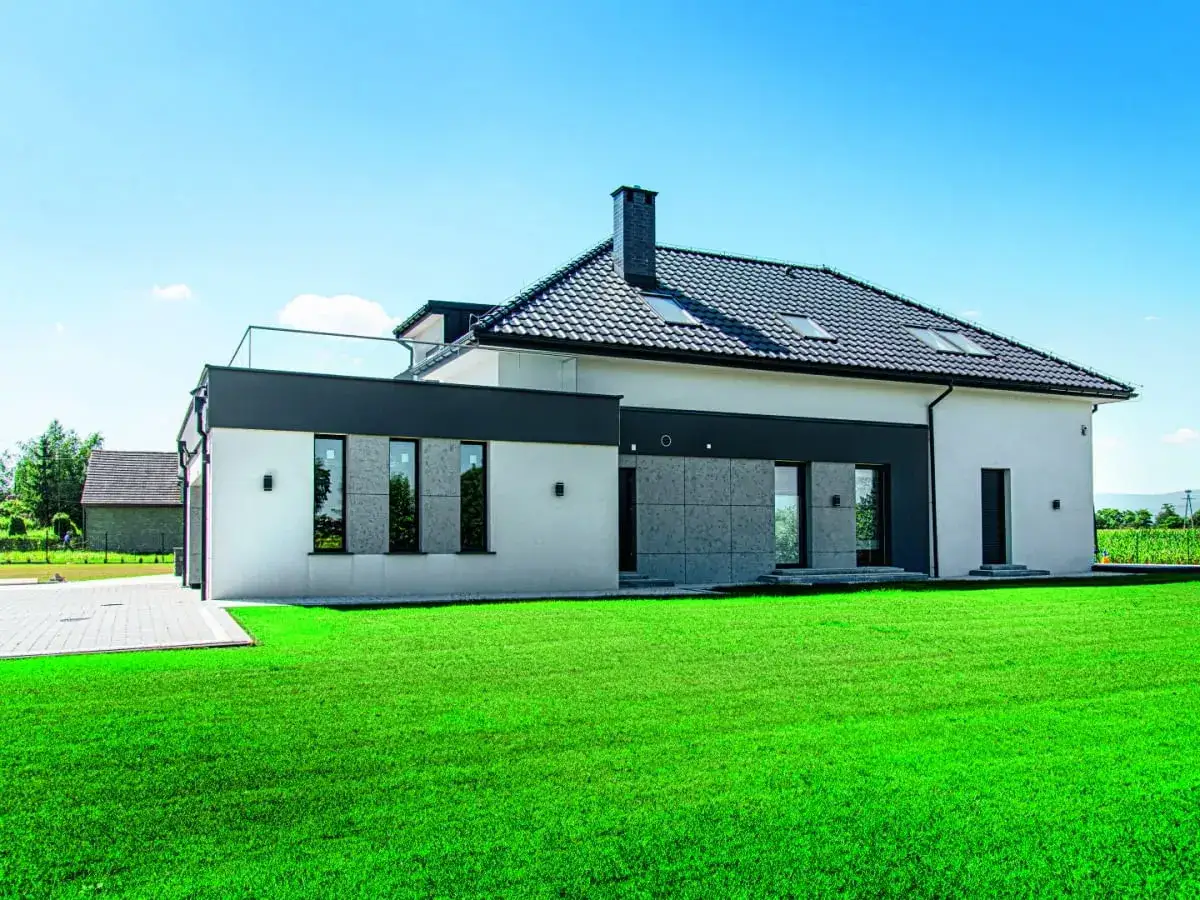 Nowoczesny dom z białym tynkiem do elewacji, szarymi panelami i dużymi oknami, otoczony zielonym trawnikiem.