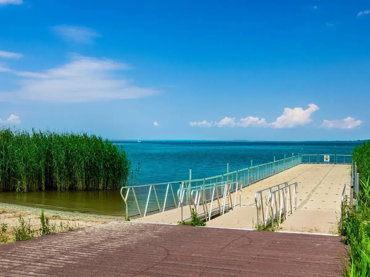 Pomost nad spokojnym jeziorem, gdzie trzciny szumią, a w oddali widać żaglówkę. Idealne miejsce, by zastanowić się, ile jest ze Szczecina do morza.