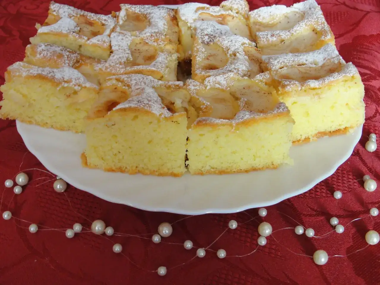 Pyszne ciasto na oleju z jabłkami, posypane cukrem pudrem, prezentuje się apetycznie na białym talerzu.