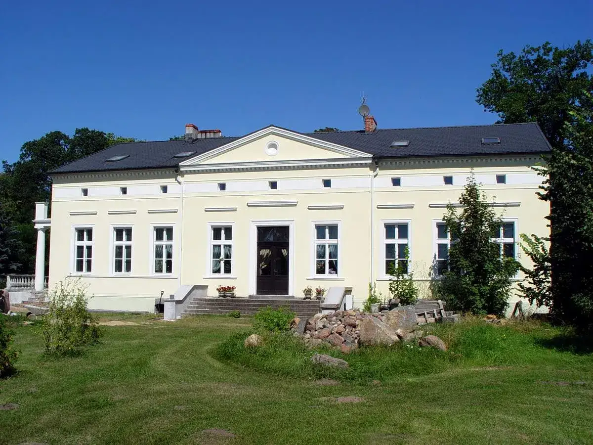 Pałac w Łącku zaprasza na zwiedzanie. Jasnoż&oacute;łty budynek z białymi zdobieniami i ciemnym dachem otoczony zielenią.