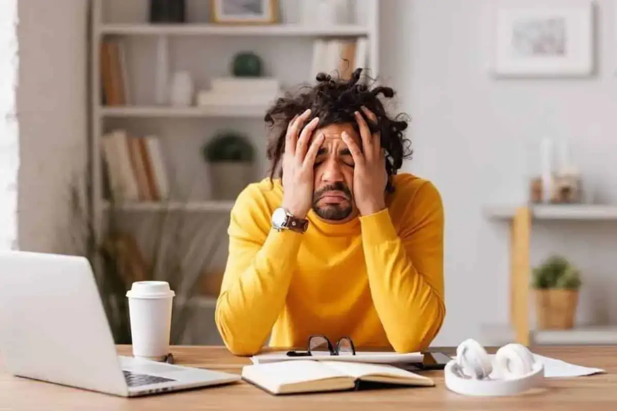 Mężczyzna z rękami na głowie, wyrażający przygnębienie. Na biurku laptop, kubek, notatnik i słuchawki. To mogą być objawy przewlekłego stresu.