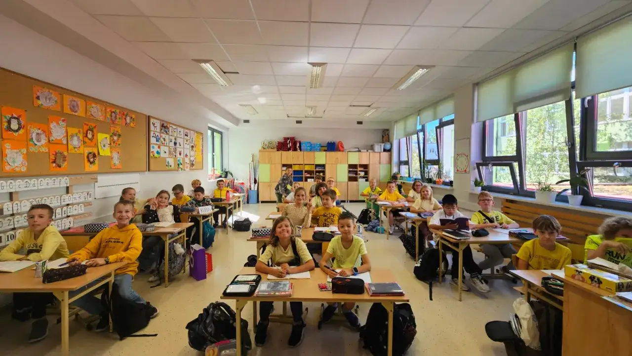Uczniowie w klasie, radośnie patrzą w obiektyw. Na ścianach wiszą prace plastyczne, świadczące o fajnych inicjatywach w szkole.