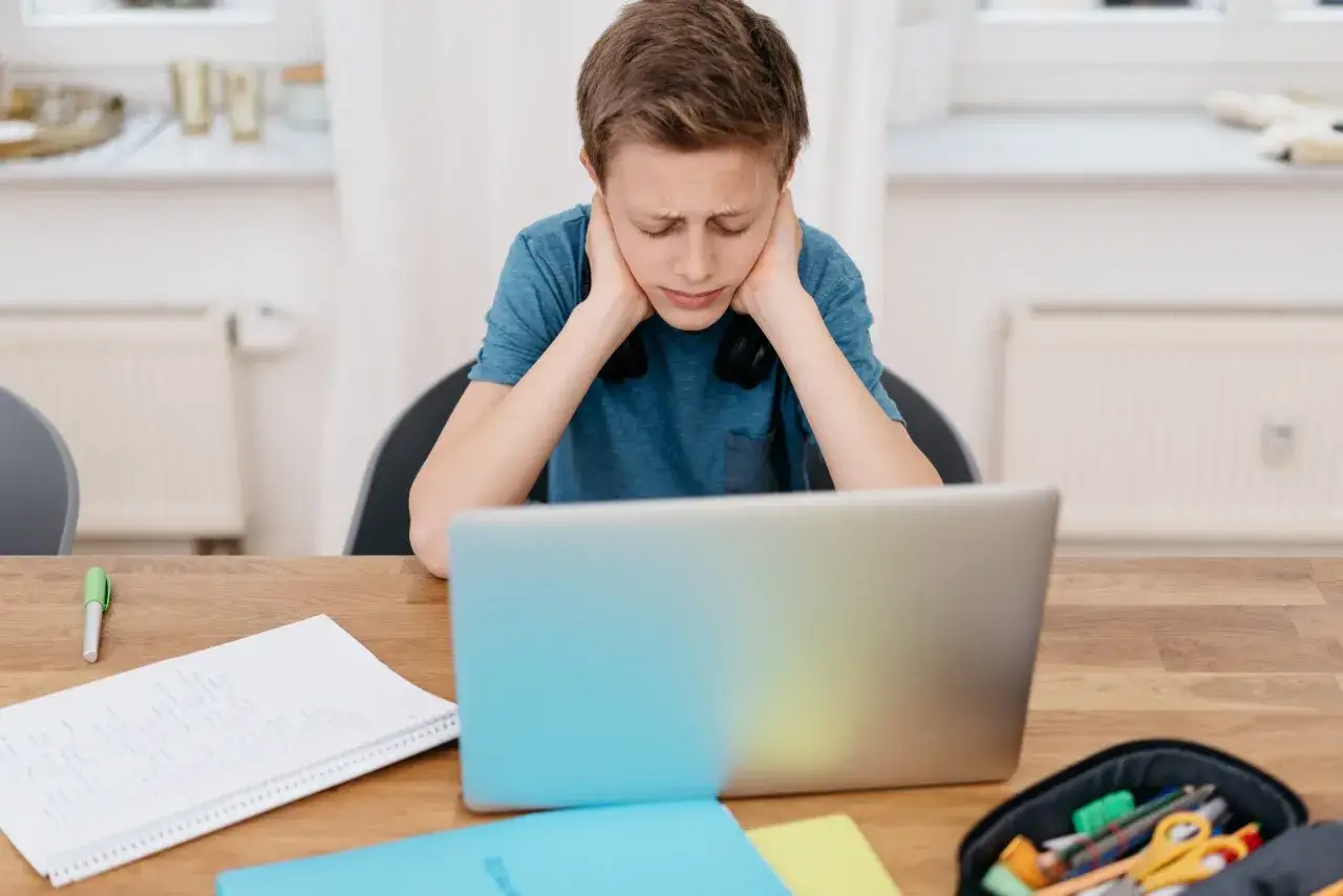 Chłopiec z rękami na skroniach przed laptopem, zmęczony długim zdalnym nauczaniem. Na biurku zeszyty i piórnik.