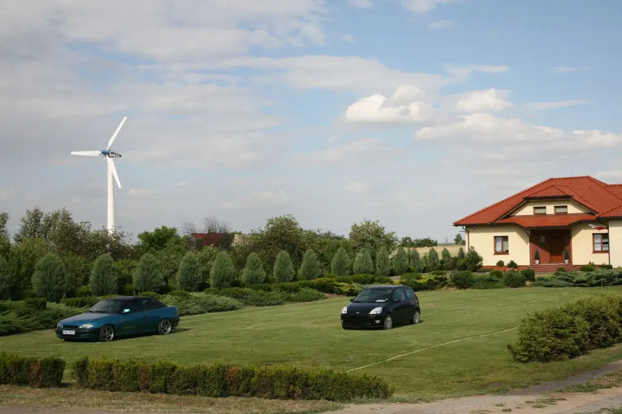 Zielony kabriolet i czarny hatchback na trawniku przed domem z czerwoną dachem. W tle biała elektrownia przydomowa.