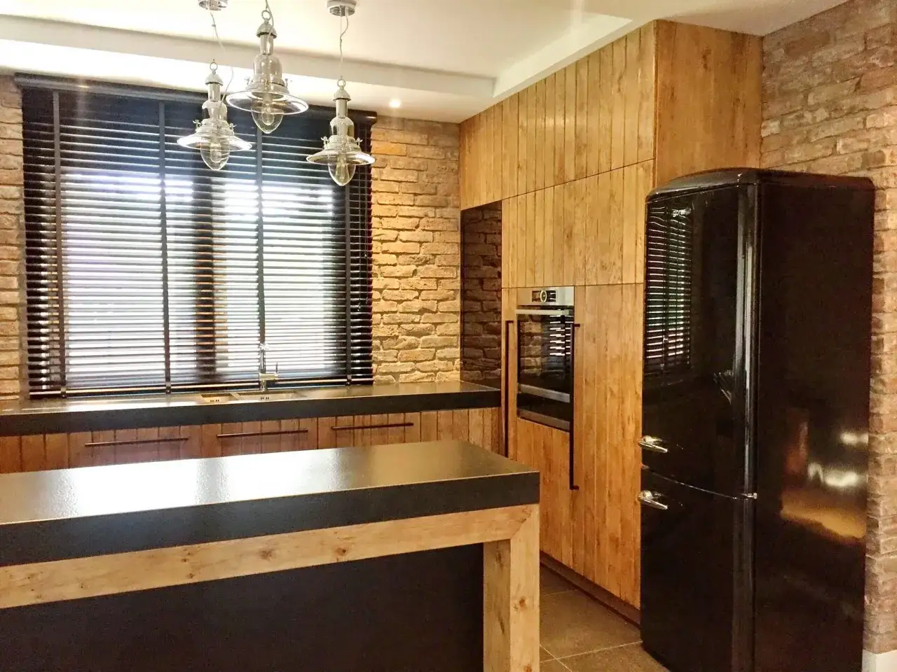 Nowoczesna kuchnia z ceglaną ścianą, drewnianymi frontami, czarną lod&oacute;wką i wyspą. Okno zasłaniają żaluzje drewniane i zasłony.