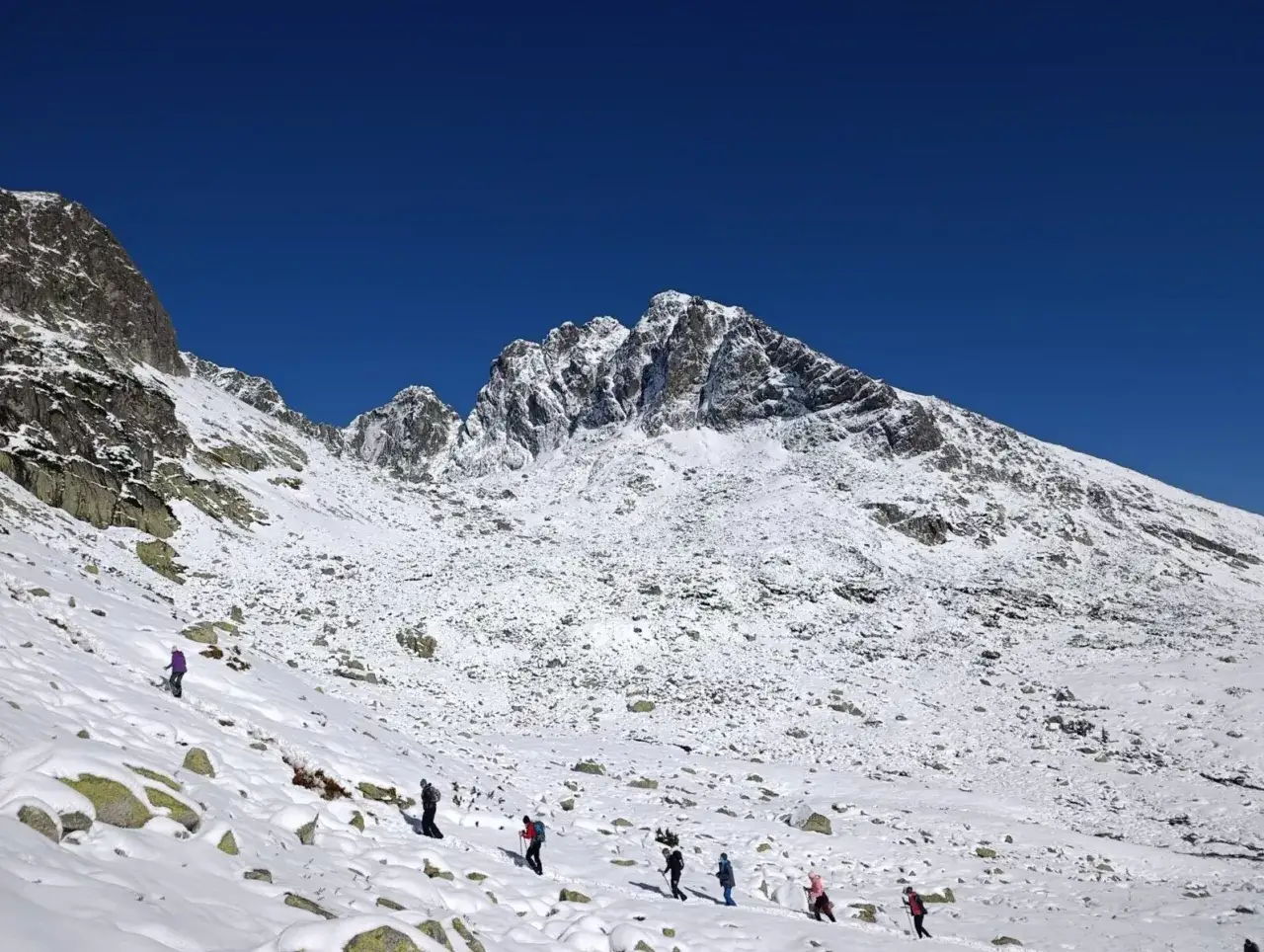 Grupa turystów wędruje po ośnieżonym zboczu. W tle majestatyczny kozi wierch zimą, pokryty śniegiem i lodem, pod błękitnym niebem.