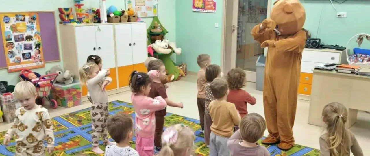 Dzieci w przedszkolu świętują dzień misia, tańcząc z pluszowym niedźwiedziem.
