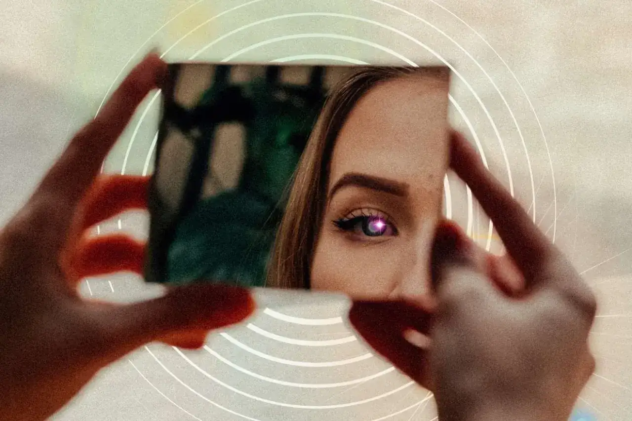 Dłoń trzyma małe lustro, w kt&oacute;rym odbija się oko kobiety z fioletową tęcz&oacute;wką. Efekt lustra tworzy surrealistyczny obraz.