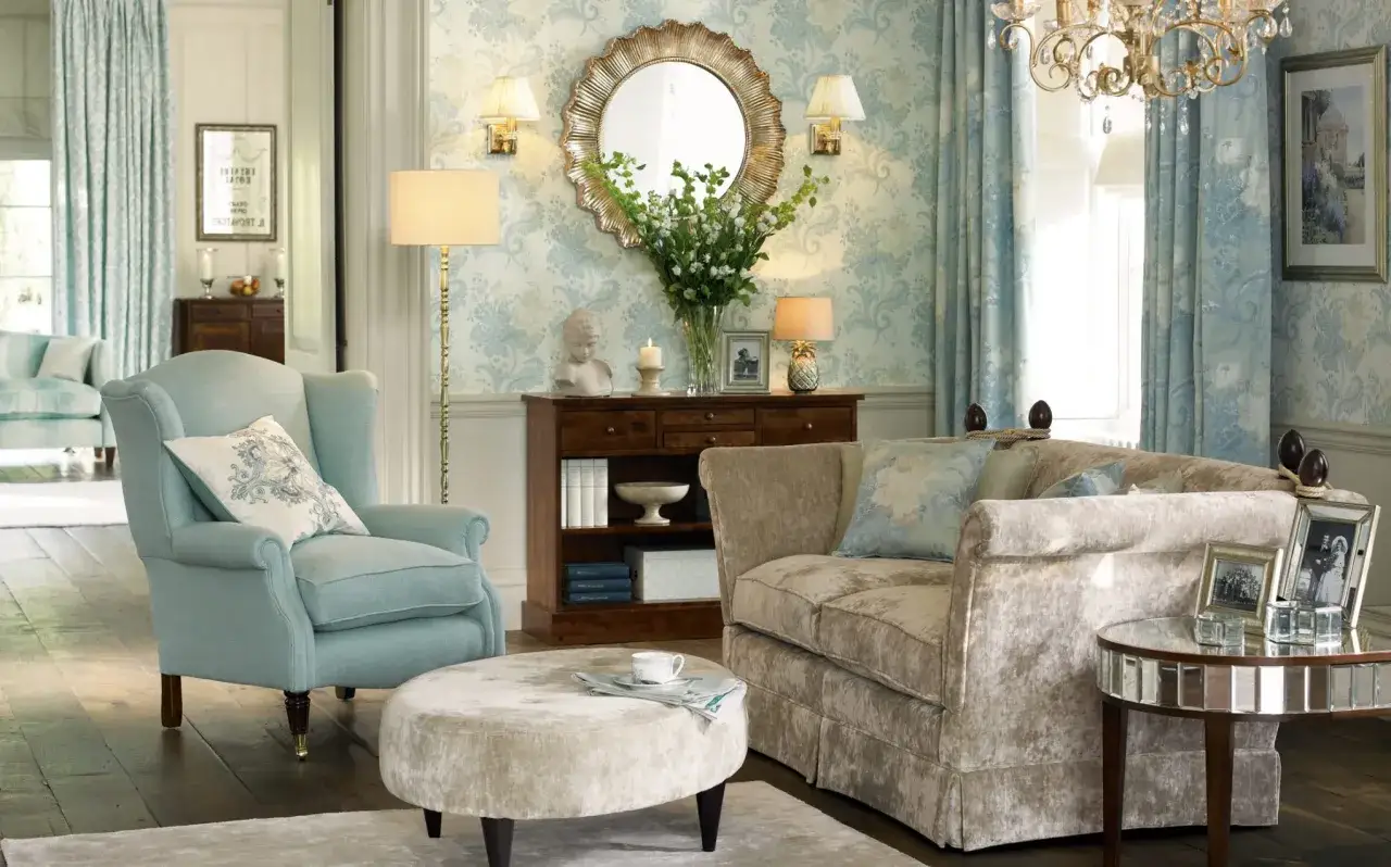 Przytulny salon w stylu angielskim: błękitny fotel, beżowa sofa, okrągła pufa, kwiatowa tapeta i kryształowy żyrandol.